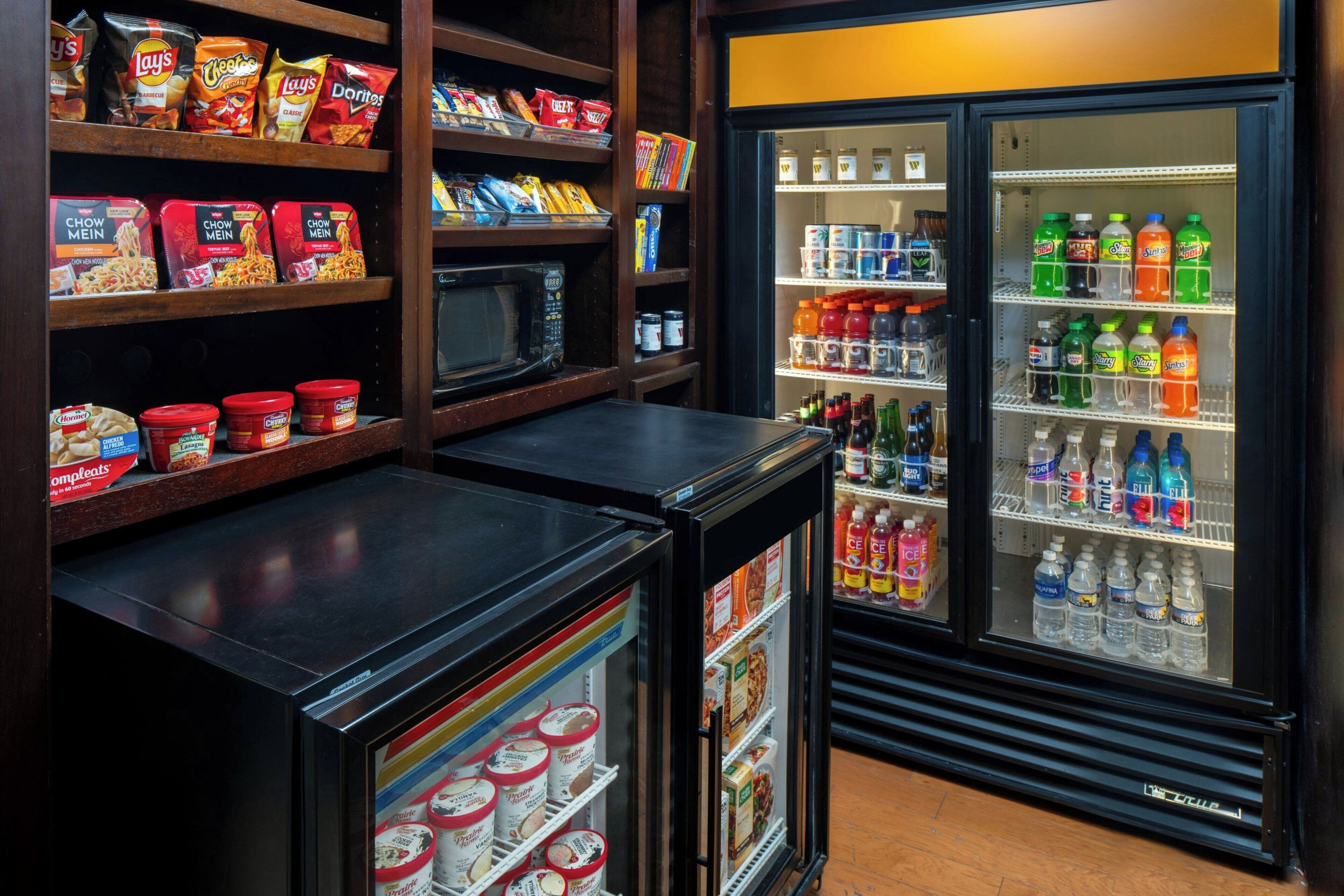 Una kitchenette con microondas, un refrigerador lleno de bebidas y estantes con aperitivos y condimentos.