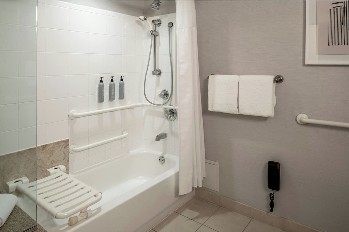 A bathroom with a bathtub, shower, grab bars, a towel rack, and a hand sanitizer dispenser.