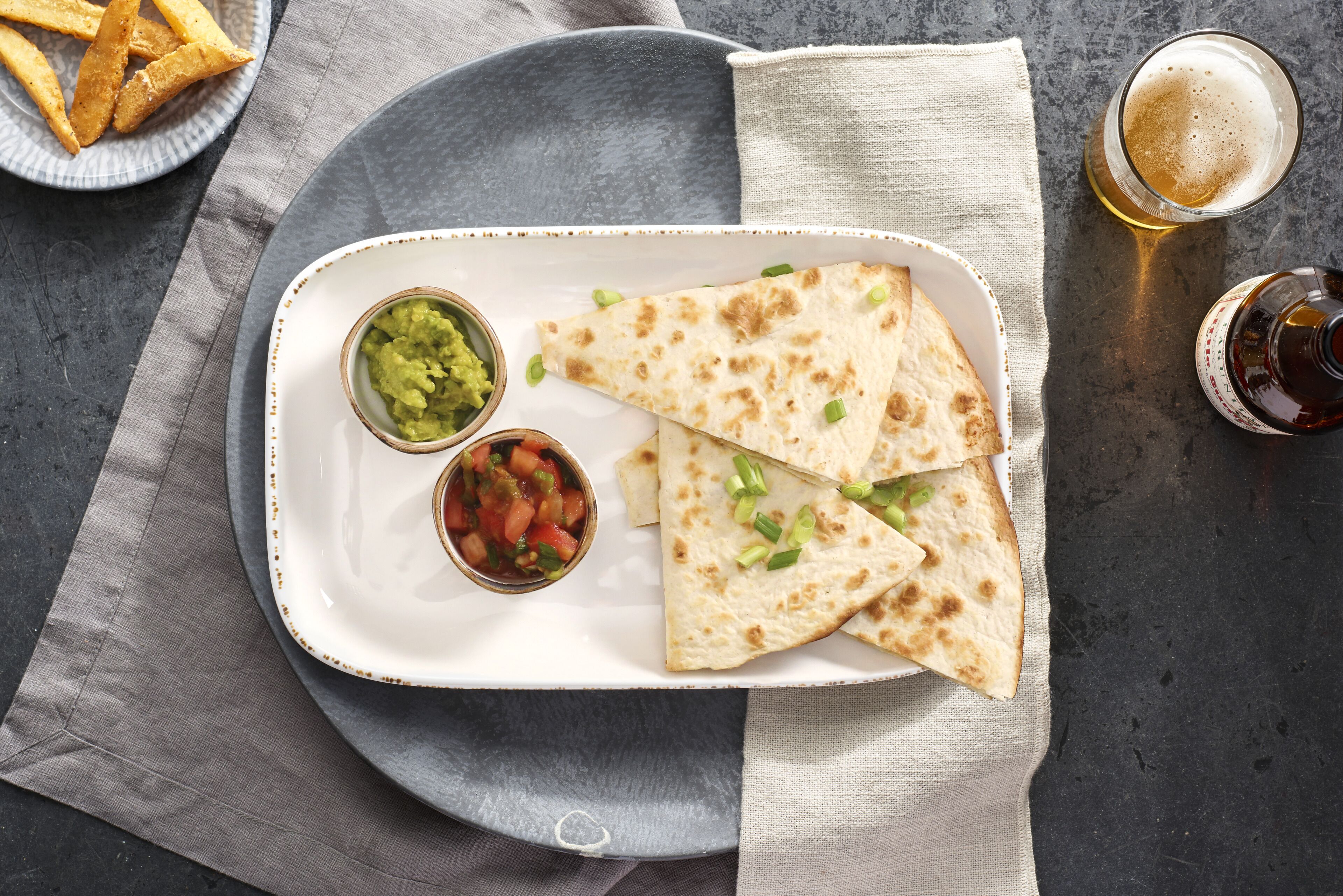 A plate with quesadillas, guacamole, and salsa, accompanied by a glass of beer and a plate of fries.