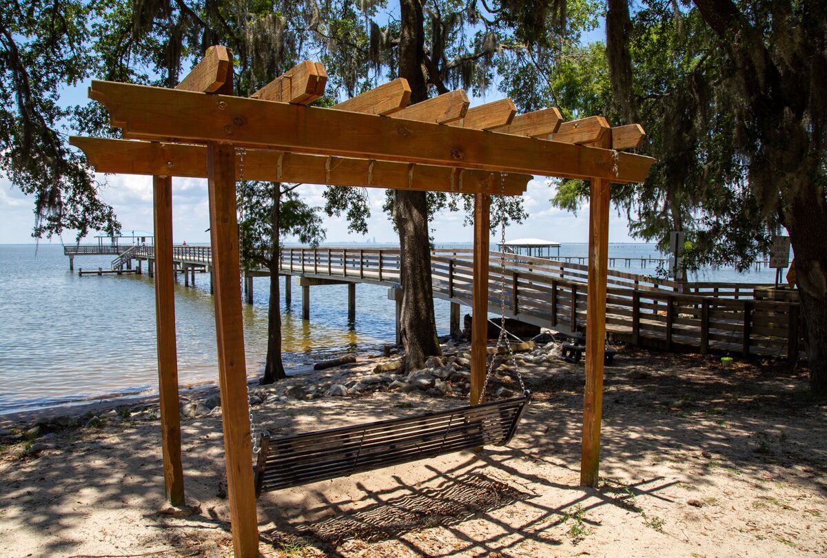 A wooden pergola with a hammock, located near a waterfront with a dock and pier in the background.