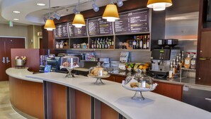 A hotel reception area with a curved counter, a menu board, and various amenities including a coffee machine, a fruit display, and a beverage dispenser. - Courtyard by Marriott Mobile Daphne/Eastern Shore (Spanish Fort)