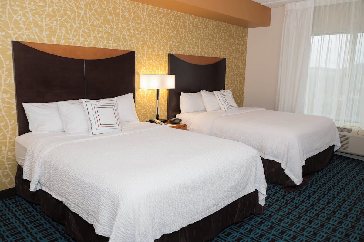 Habitación de hotel con dos camas, una mesita de noche con lámpara y una alfombra estampada.