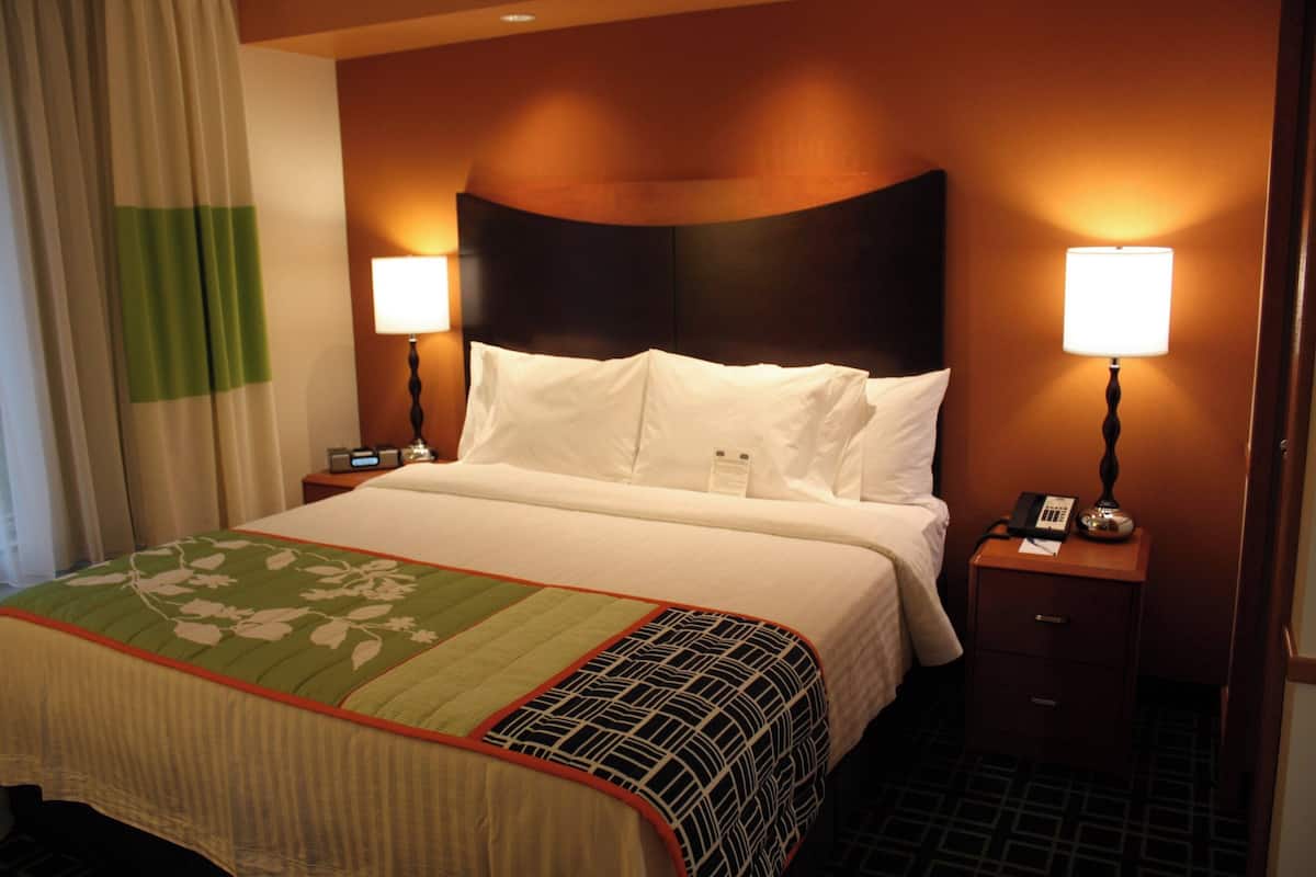 Una habitación de hotel con una cama, dos lámparas de noche, una mesita de noche con teléfono y una colcha de cama con estampado floral en verde y blanco.