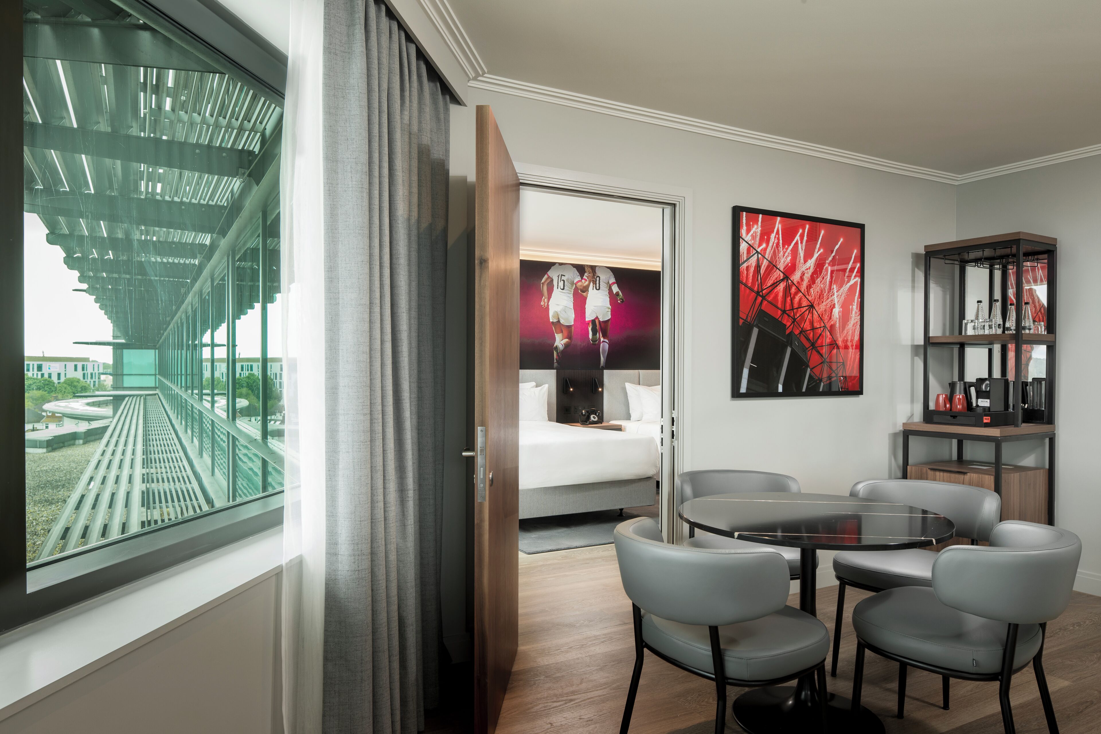 A modern hotel room with a large window, a bed, a round table with chairs, and a bookshelf.