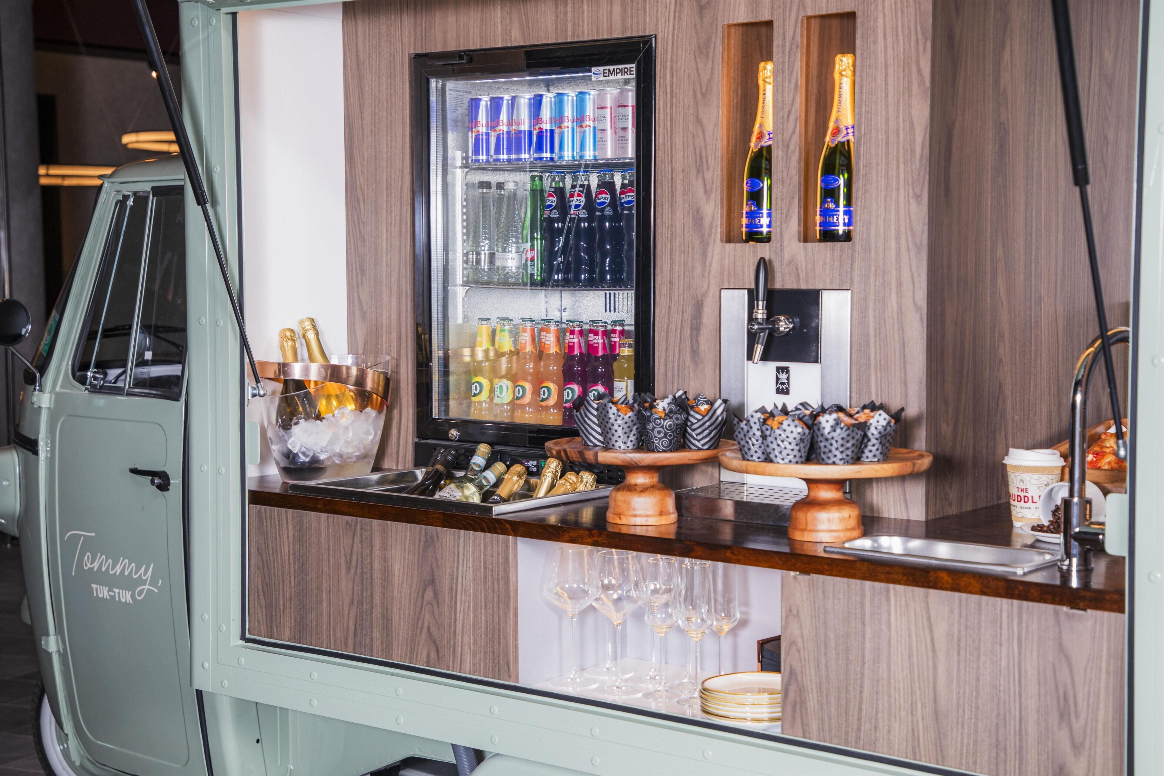Eine Barbereich mit einem Kühlschrank, der mit Getränken gefüllt ist, einer Champagner-Ausstellung und einer Theke mit Gläsern und einer Kaffeetasse.