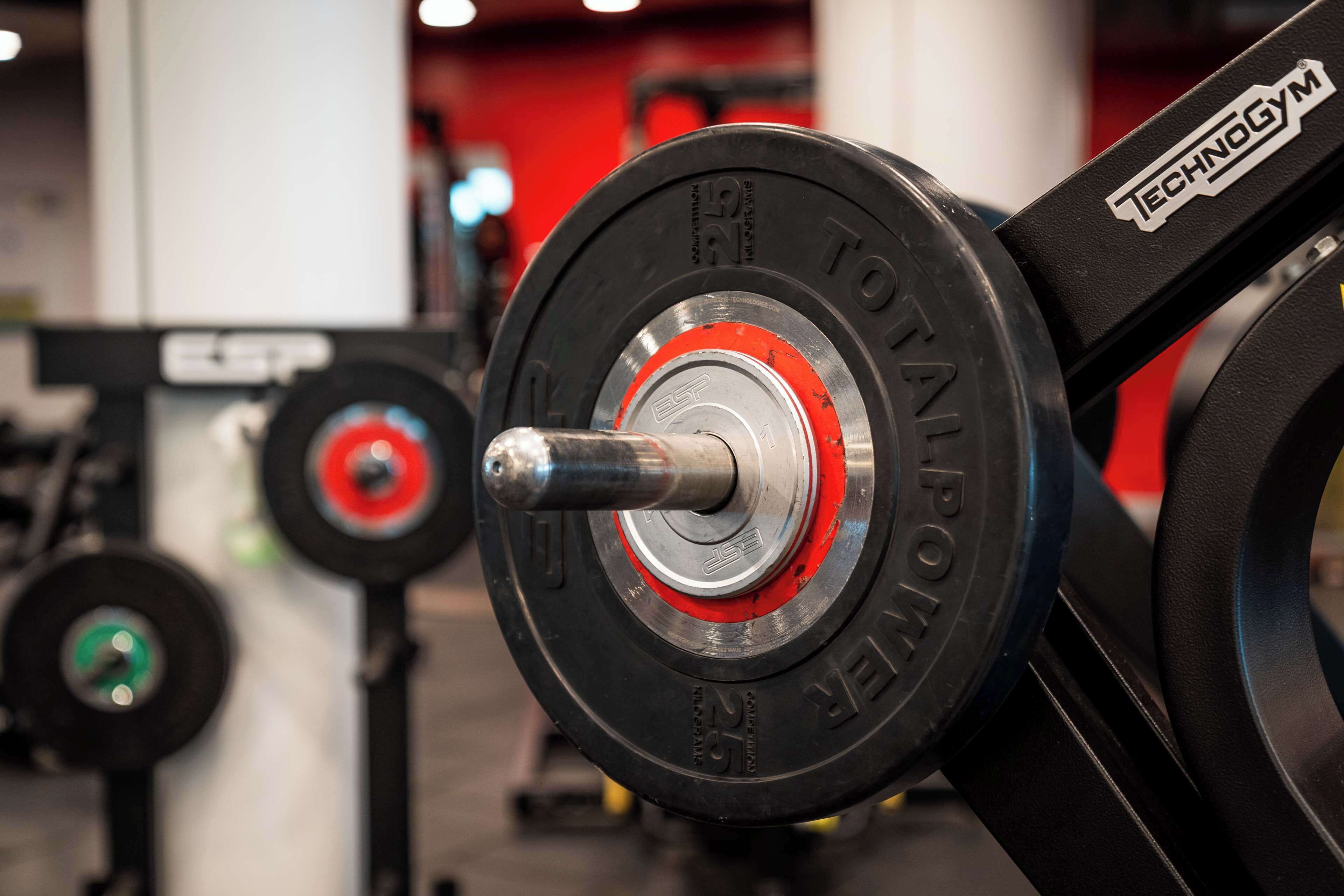 Une barre d’haltérophilie avec des disques de poids rouges et noirs, une barre argentée et une étiquette indiquant « TECHNOGYM ».