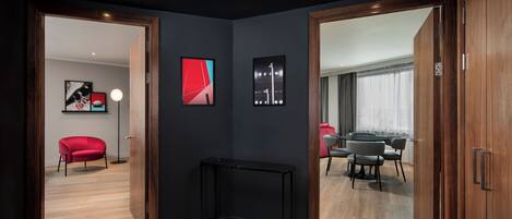 A modern hotel room with a dark wall, a wooden floor, a black console table, and a dining area with a gray sofa and a red chair.