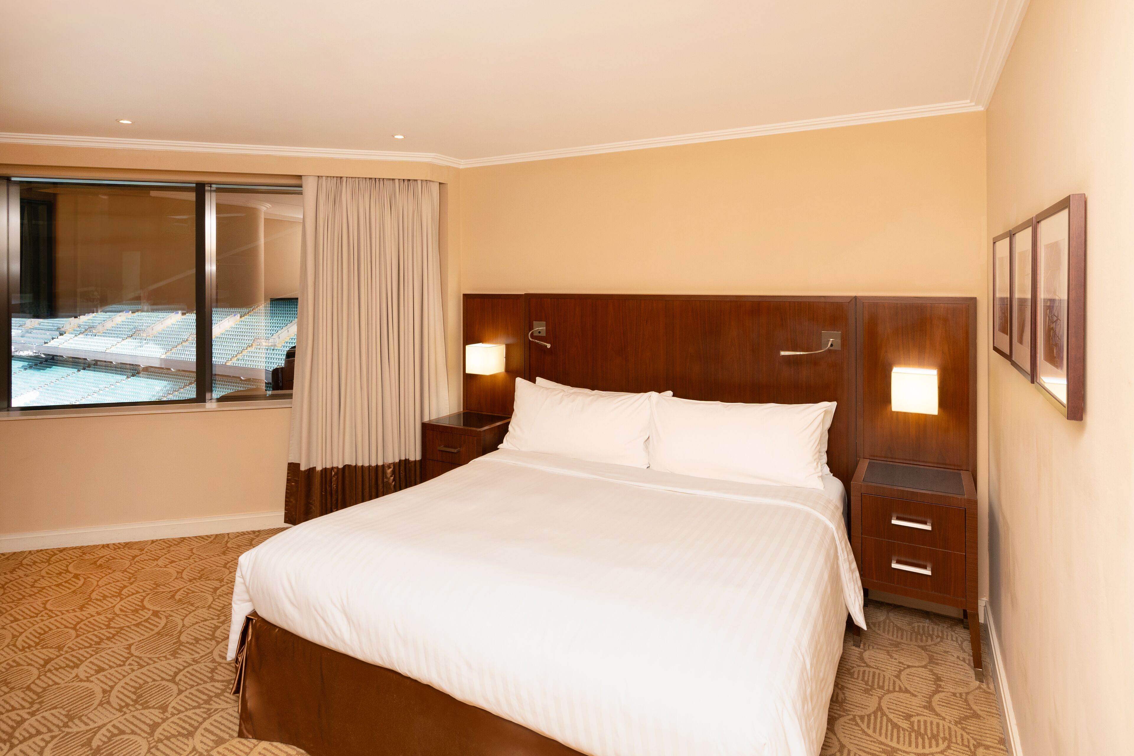 A hotel room with a large bed, wooden headboard, two bedside tables, a window with curtains, and a view of a pool area.