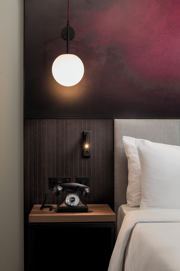 A modern hotel room with a wooden nightstand, a classic rotary phone, a wall-mounted light fixture, and a bed with white bedding.