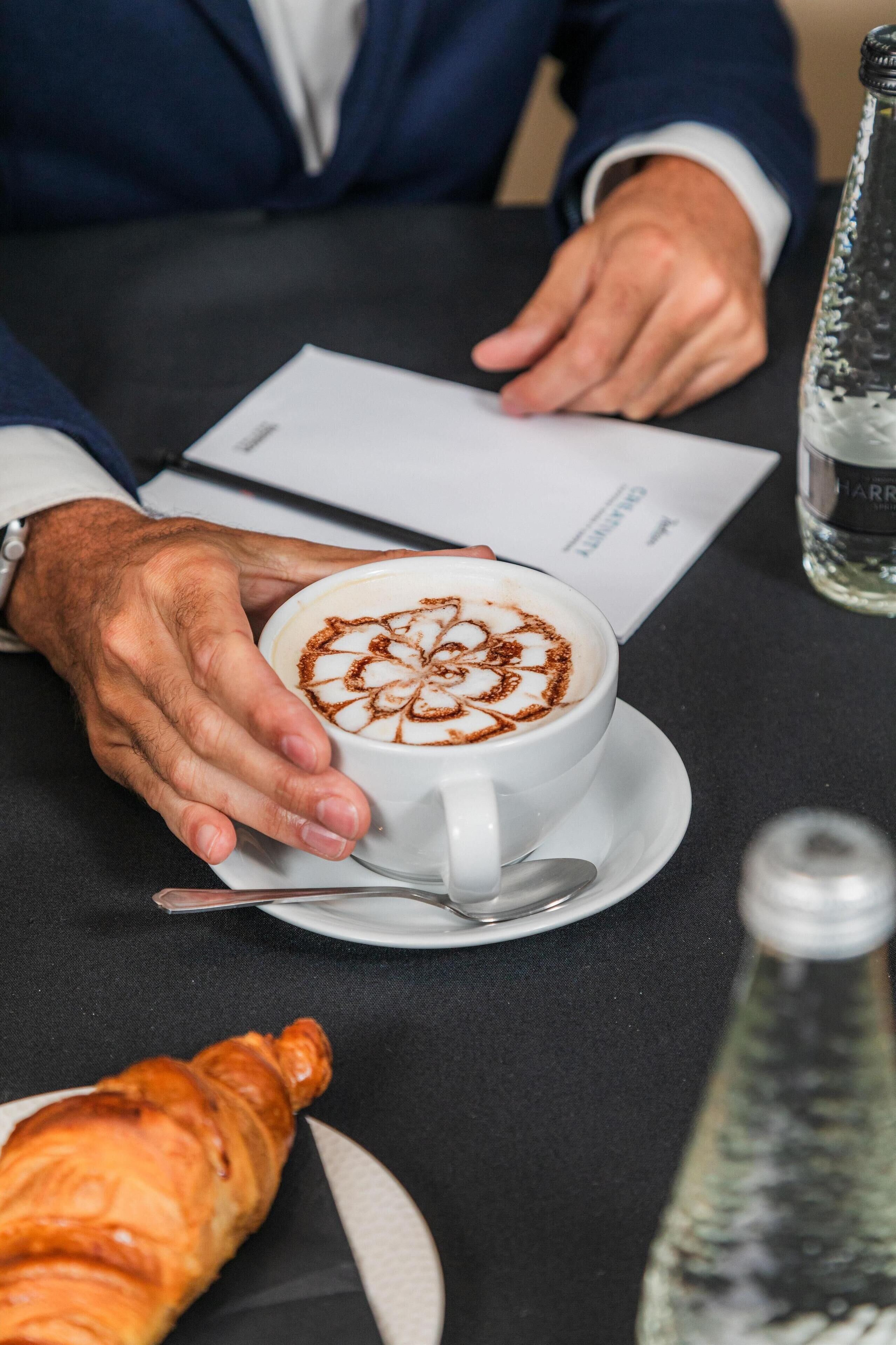 Een persoon houdt een koffiekopje met latte-kunst vast, met een croissant en een fles water op een tafel.