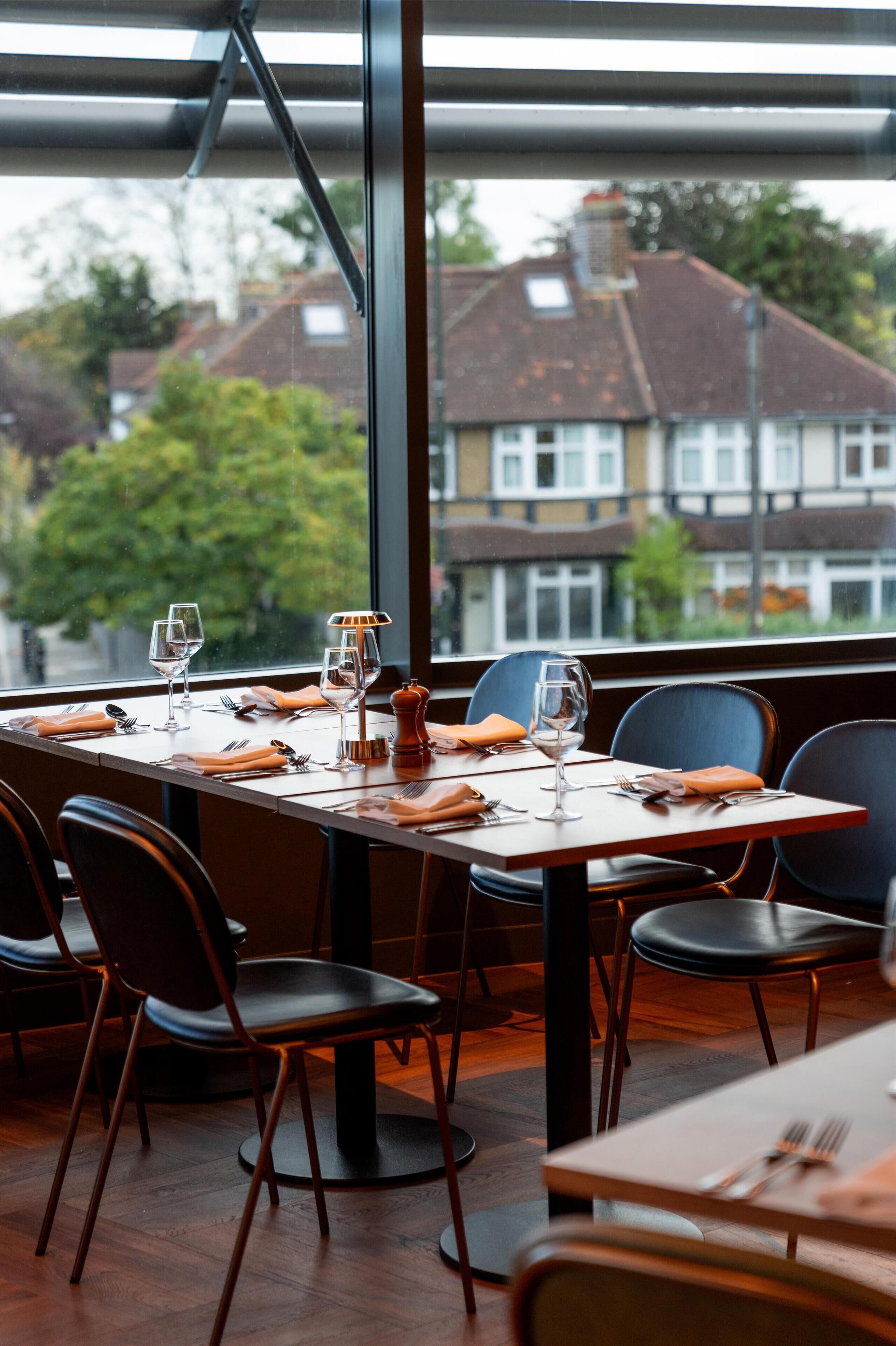 A dining area with a table set for two, chairs, and a view of buildings through large windows.