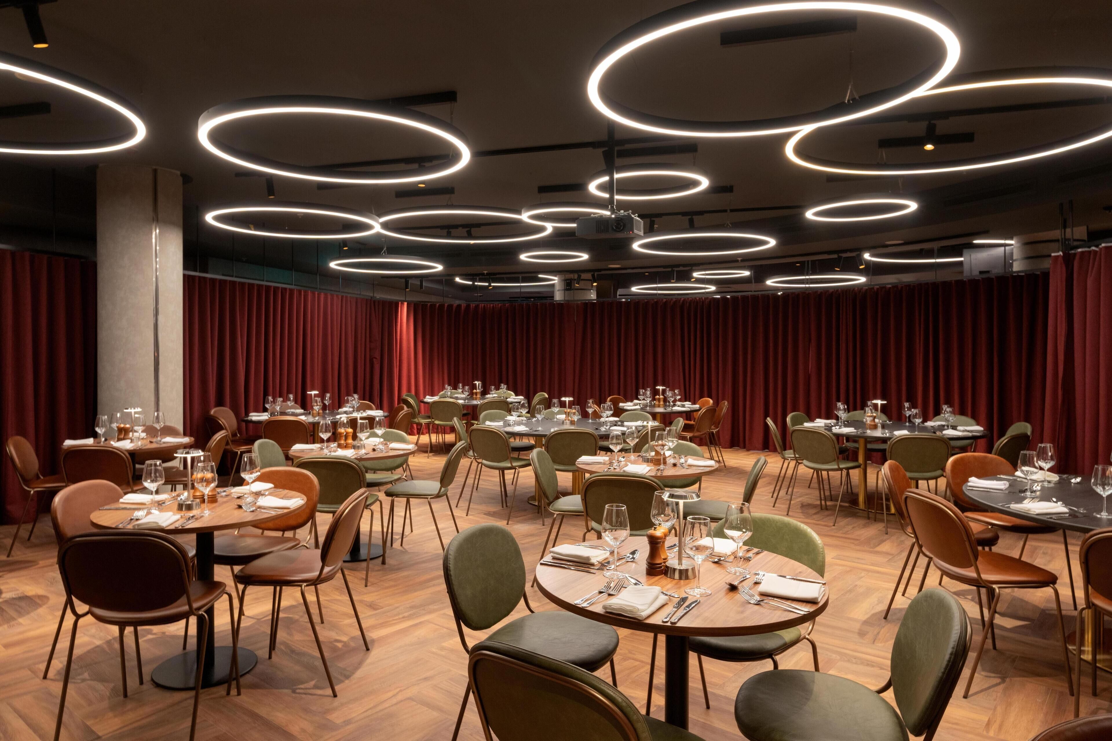 A dining area with multiple round tables, each set with plates, cutlery, and glasses. The room features a modern ceiling with circular light fixtures and red curtains along the walls.