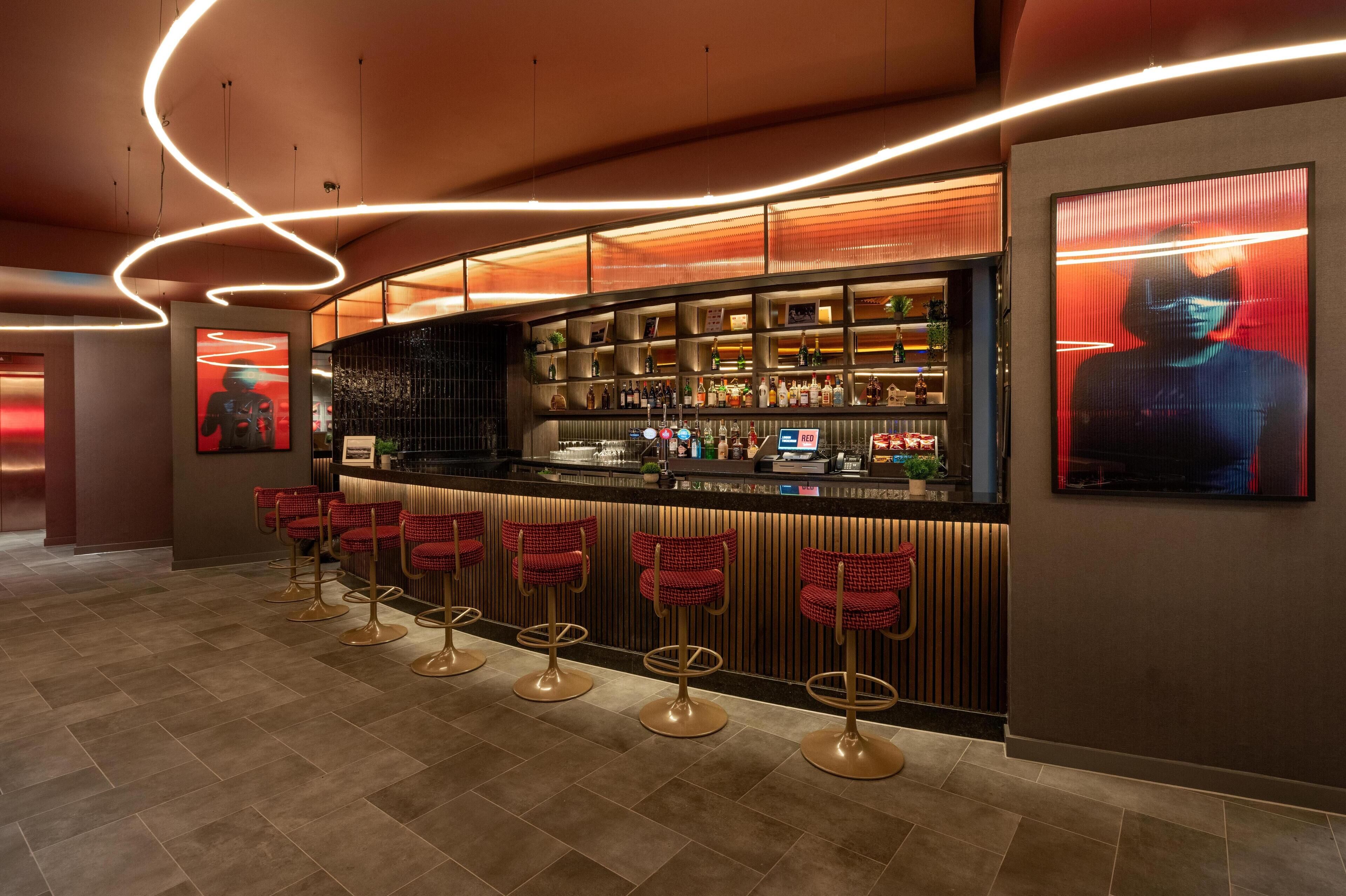 Una zona de bar con un mostrador curvo, varias estanterías llenas de botellas y taburetes rojos.