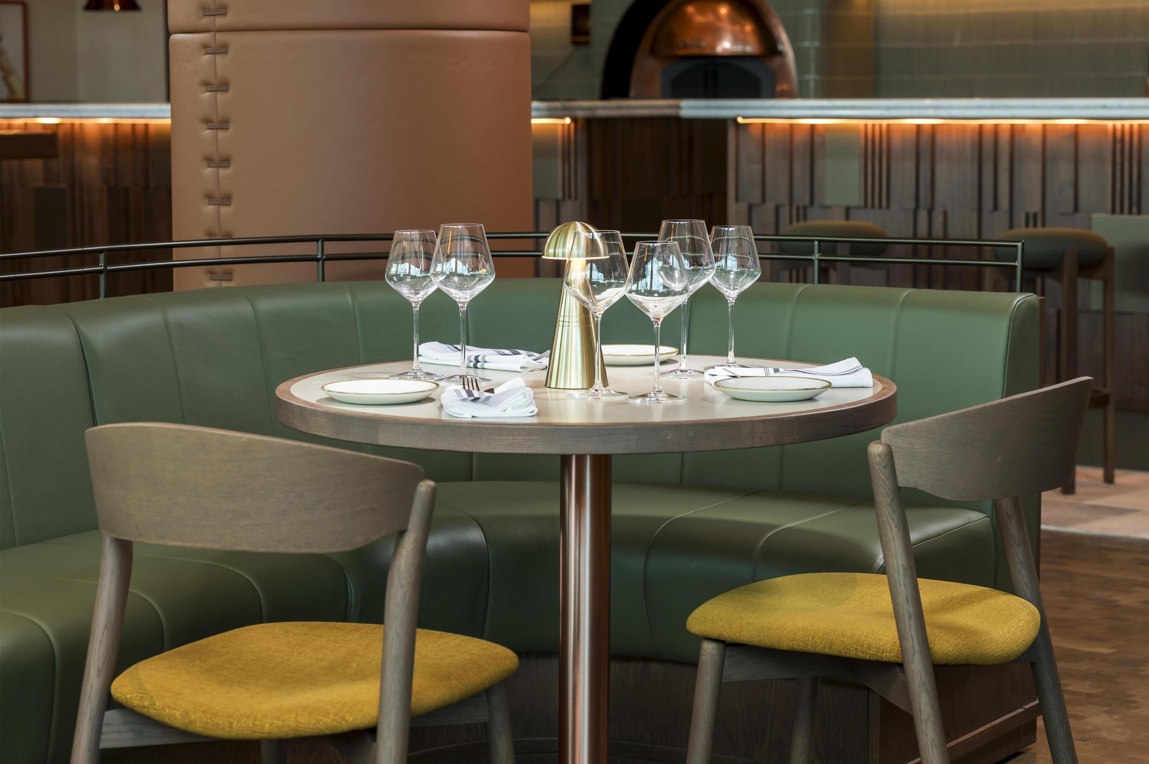 Un restaurante con mesa redonda preparada para dos personas, asientos tipo cabina tapizados en verde y una decoración de estilo moderno.