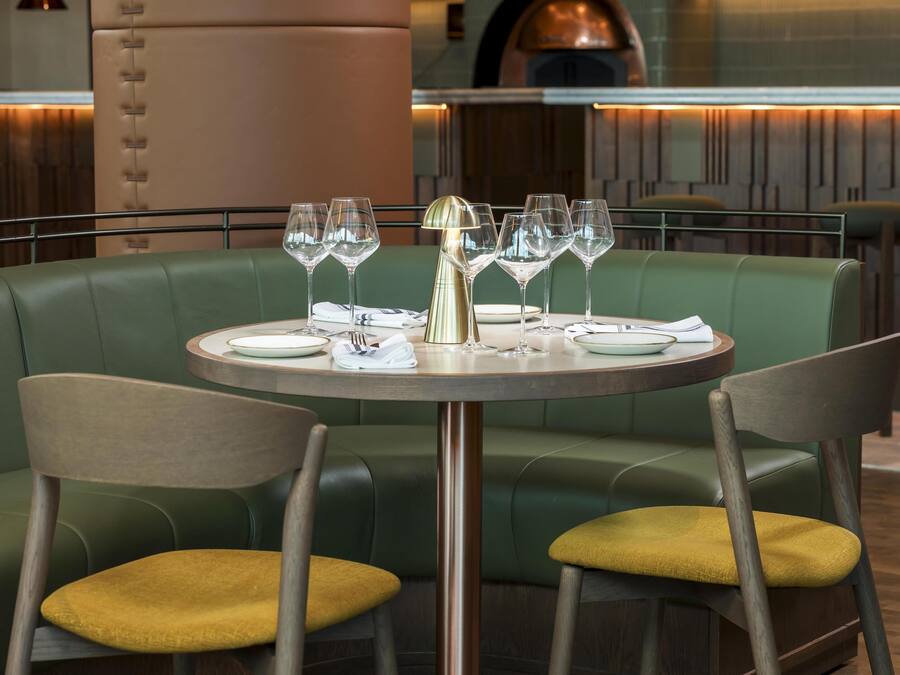 Un restaurante con mesa redonda preparada para dos personas, asientos tipo cabina tapizados en verde y una decoración de estilo moderno.