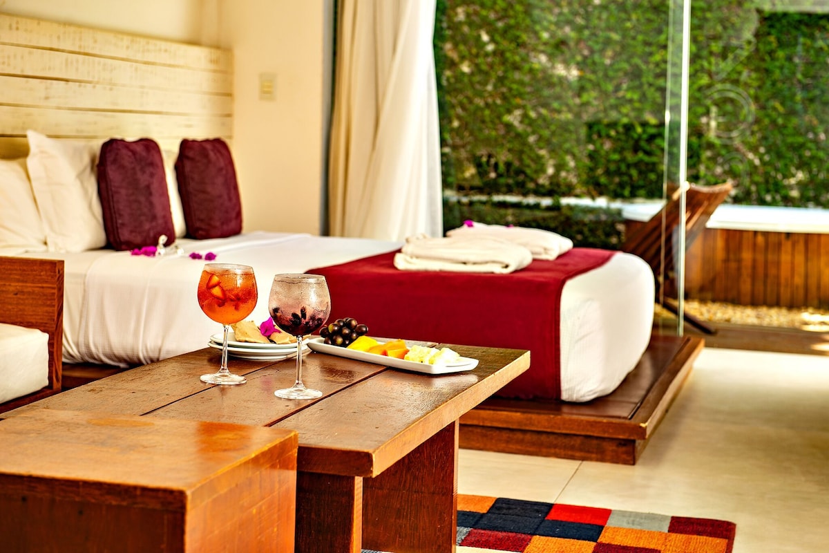 A hotel room with a bed, a wooden table with drinks and fruit, and a view of greenery outside.
