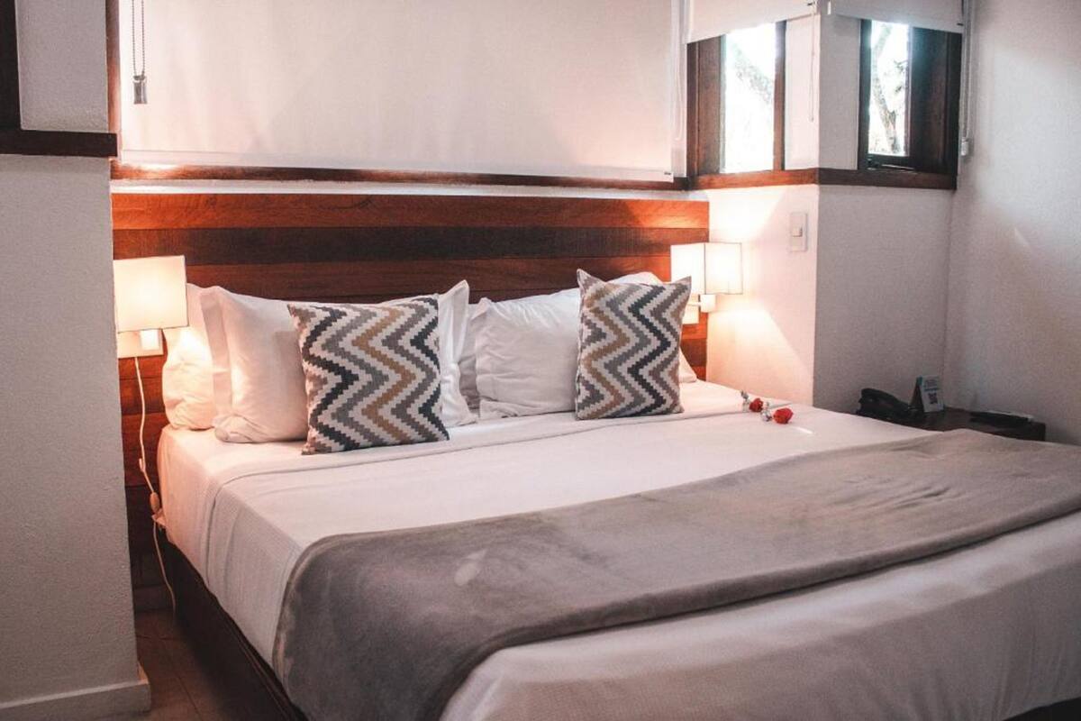 Uma cama bem arrumada com cabeceira de madeira, roupa de cama branca e almofadas estampadas.