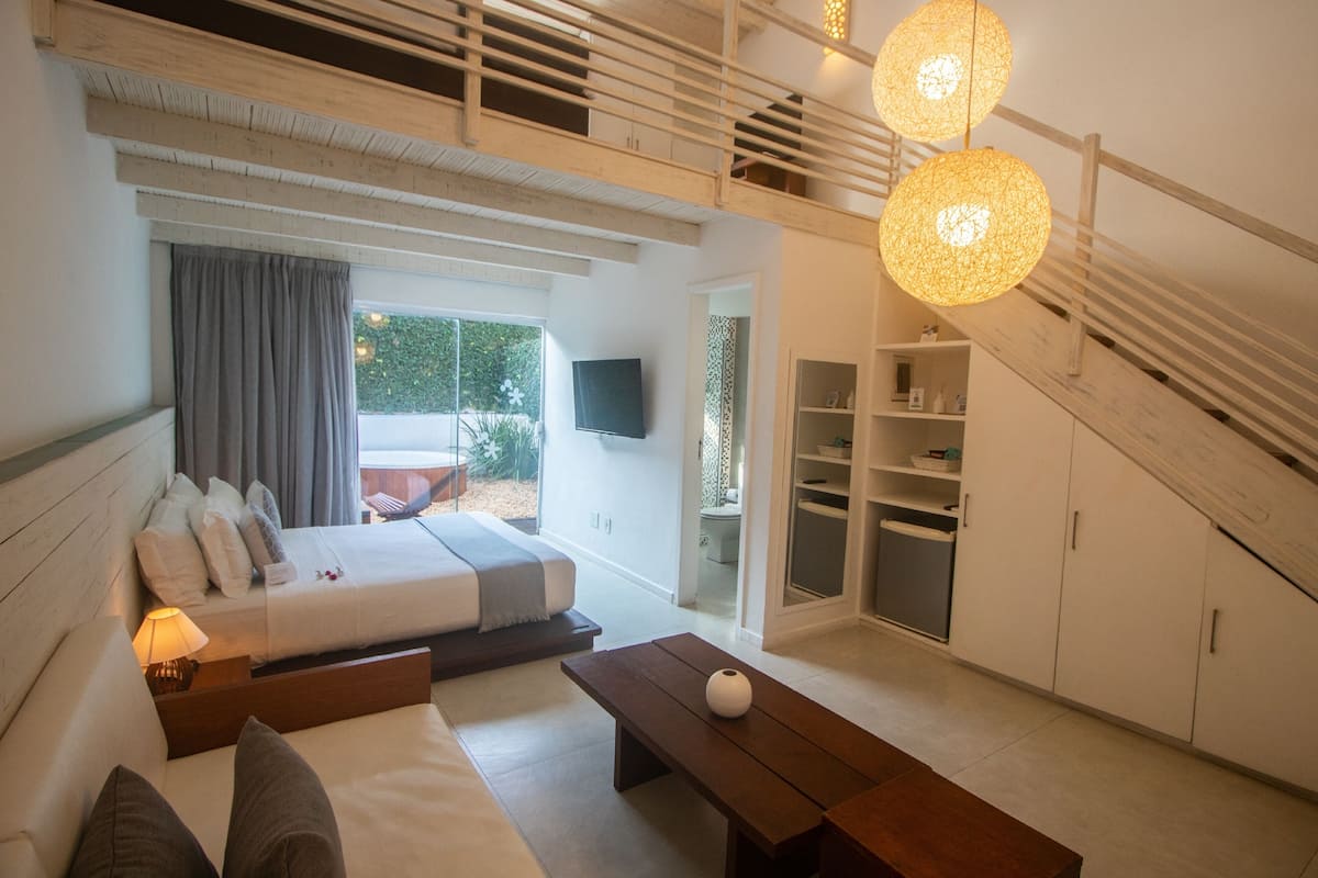 Quarto moderno com cama, sofá, um banco de madeira e uma televisão. Existe uma escada que leva a uma área externa.