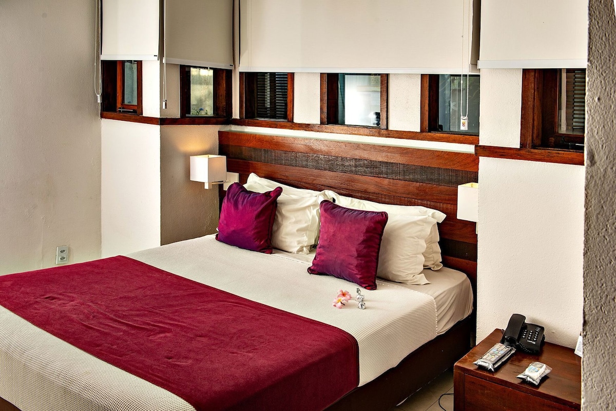 A hotel room with a bed, a nightstand with a phone and a remote, and a wall-mounted lamp.