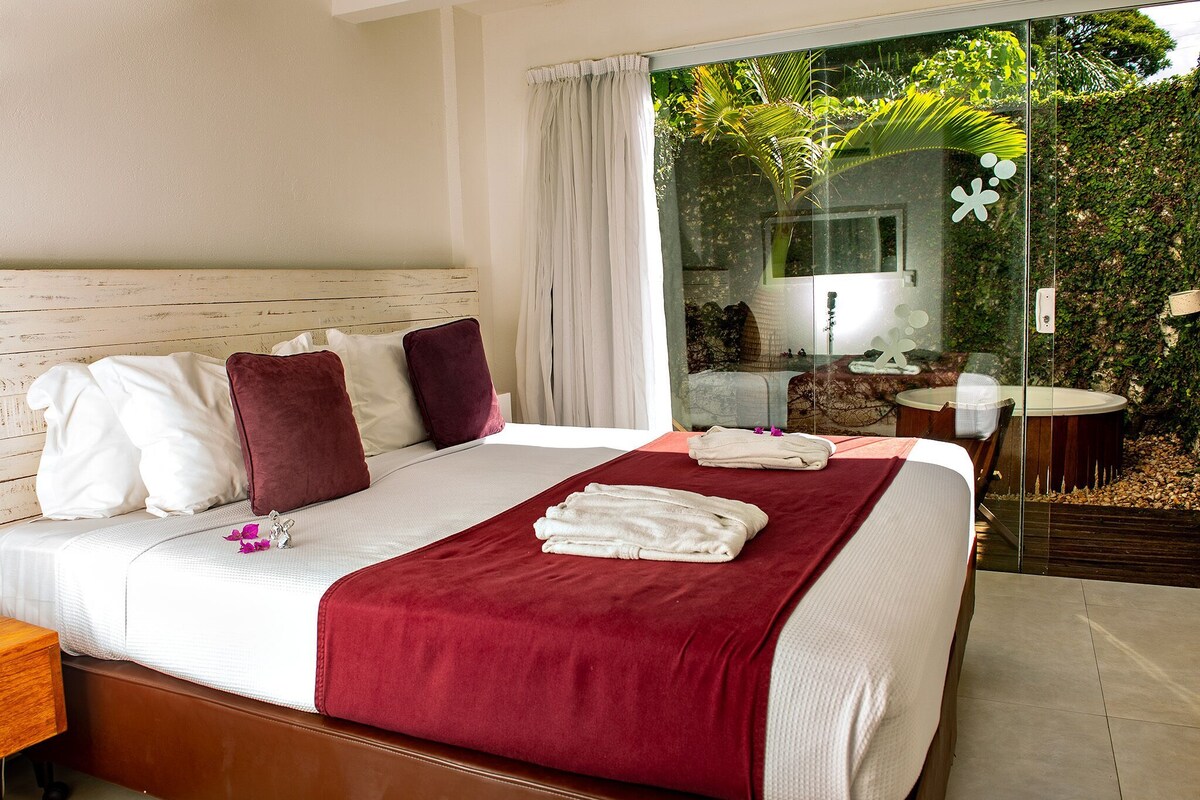 A hotel room with a bed, a nightstand, a window with curtains, and a view of greenery outside.
