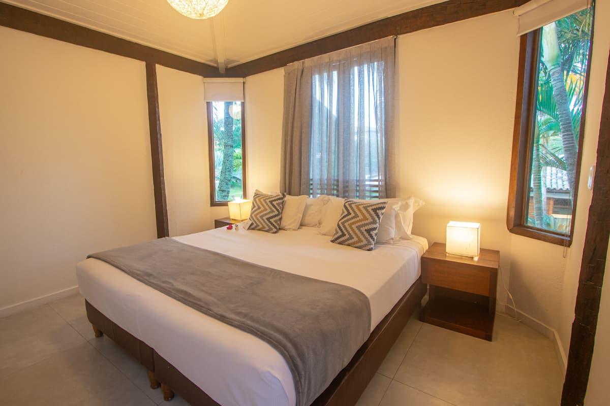 Quarto com uma cama grande, cabeceira de madeira, duas mesas de cabeceira com abajures e uma janela com cortinas.