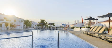 A swimming pool with lounge chairs and umbrellas, surrounded by buildings with white facades and palm trees.