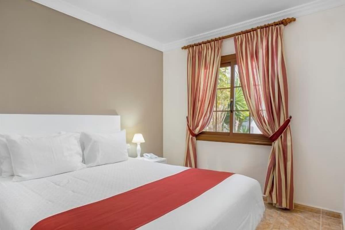 Habitació d'hotel amb llit, una taula de nit, una làmpada i una finestra amb cortines.