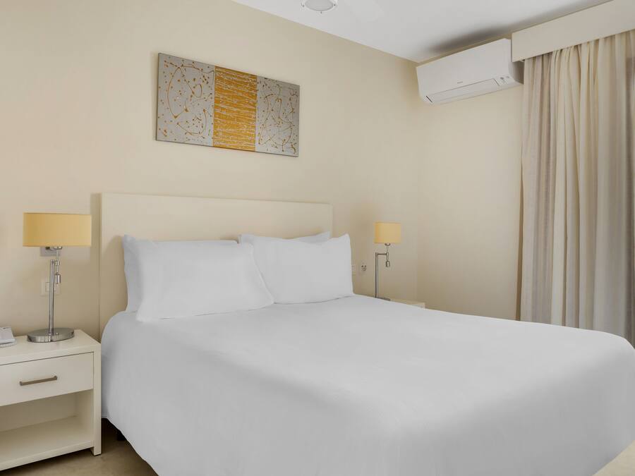 A hotel room with a large bed, two bedside tables with lamps, a wall-mounted air conditioner, and a window with curtains.