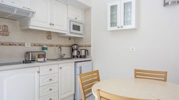 Cucina compatta con mobili bianchi, forno a microonde, macchina da caffè e frigorifero. Un tavolo da pranzo in legno con sedie.