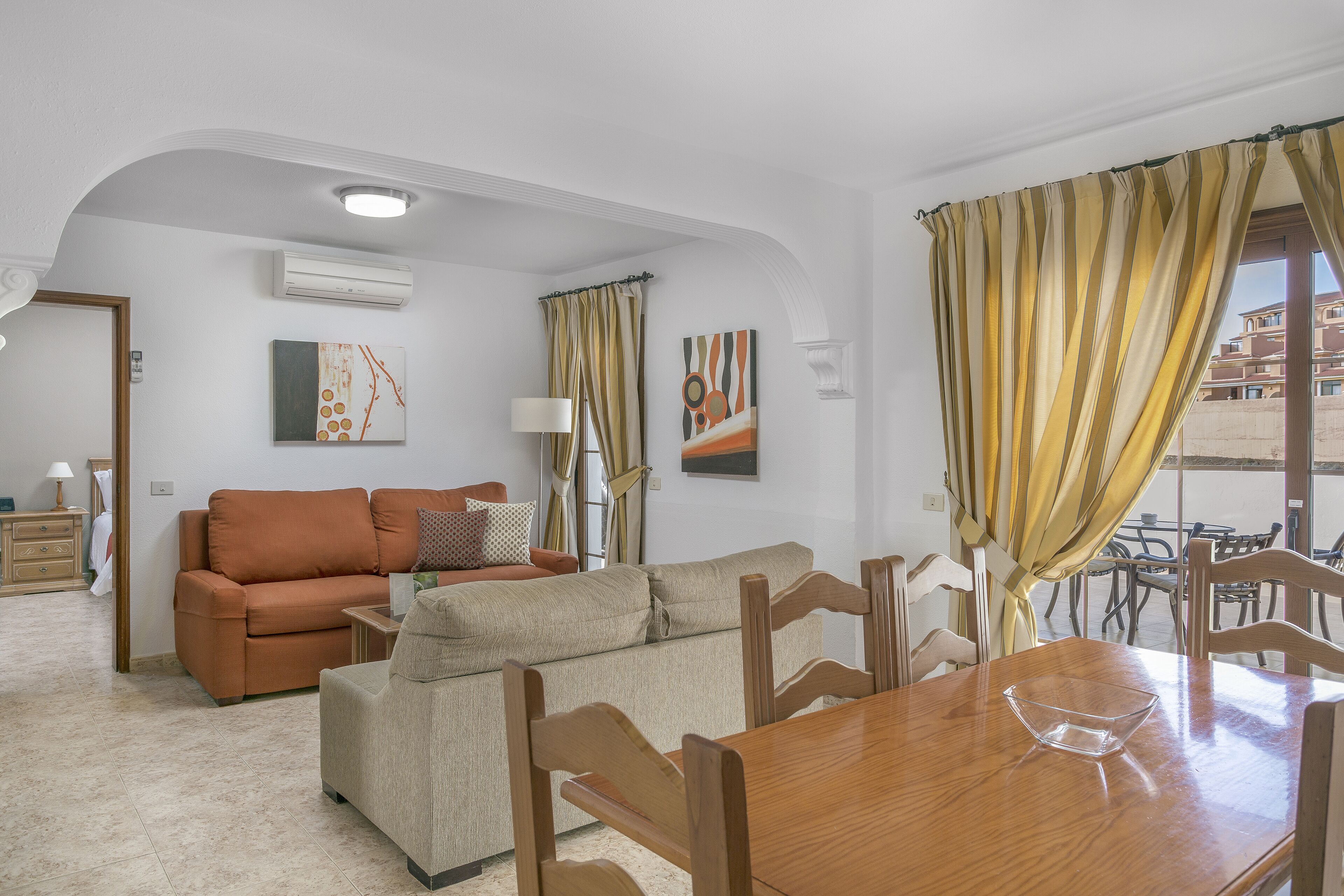 Una sala de estar con un sofá, una mesa de comedor de madera y sillas. Hay una ventana con cortinas, un equipo de aire acondicionado y obras de arte en las paredes.