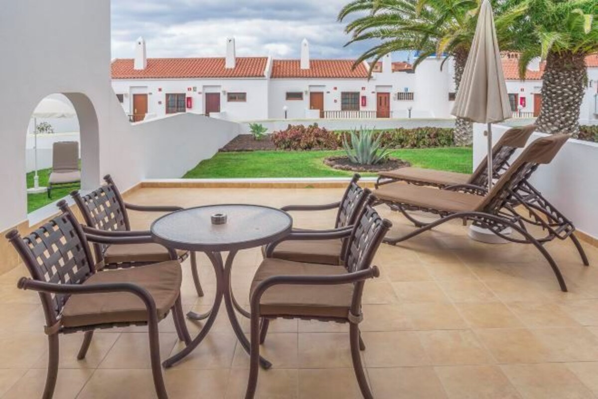 An outdoor patio with a round table and four chairs, a palm tree, and a view of a building with a red roof.