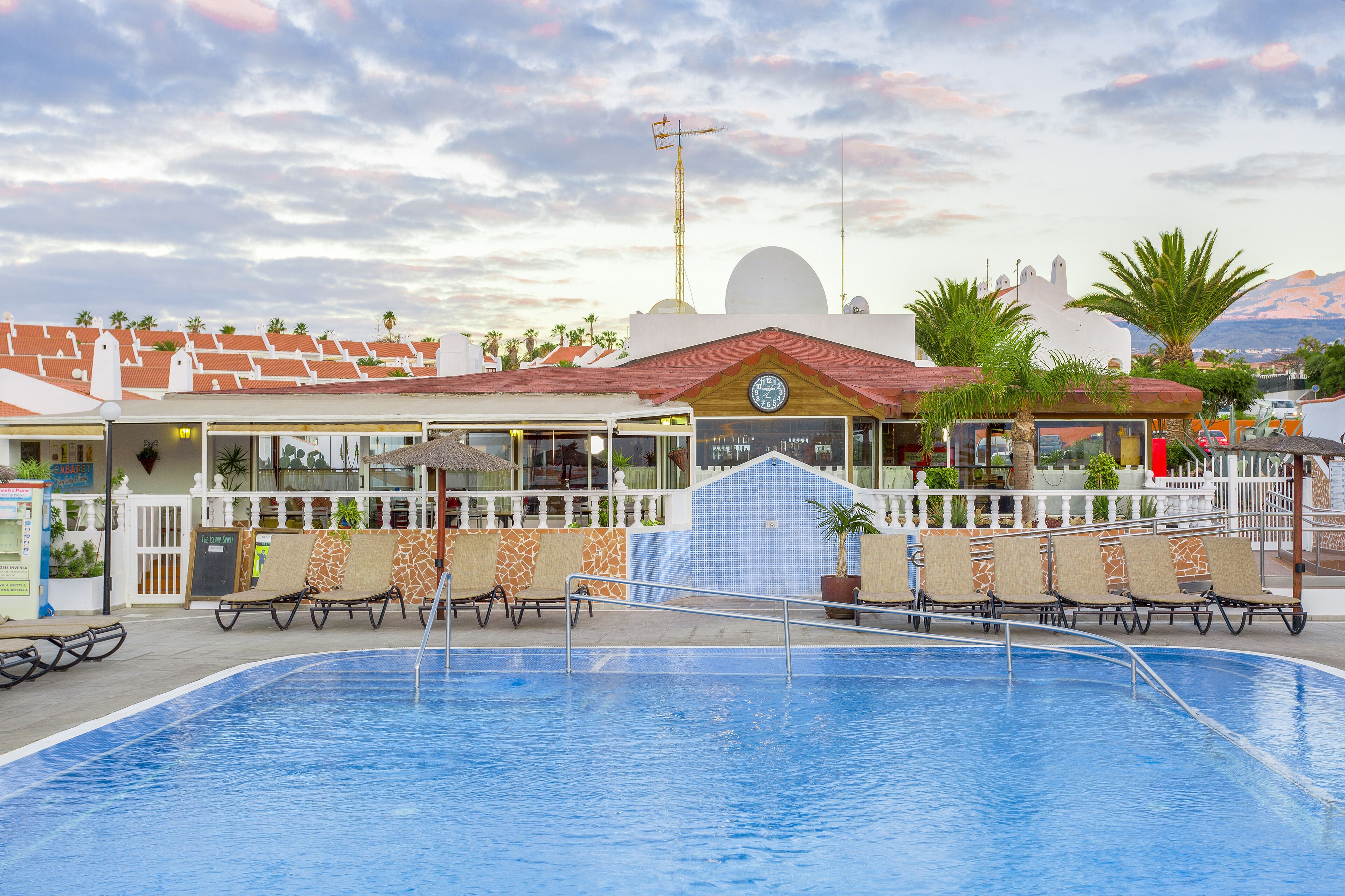 A swimming pool with lounge chairs, a bar, and a restaurant with a white fence and palm trees.