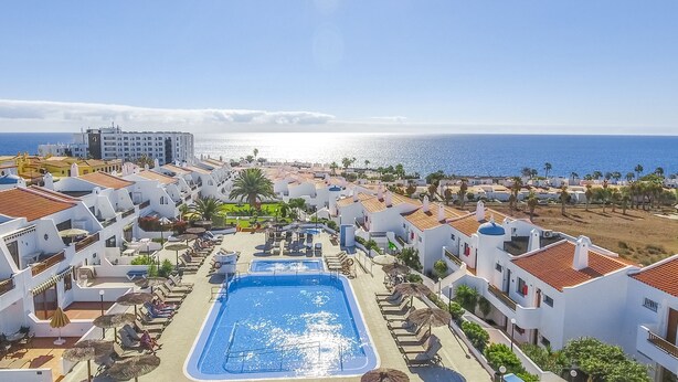 A resort with a central swimming pool, white buildings with red-tiled roofs, and a view of the sea.