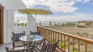A balcony with a glass table and chairs, a yellow umbrella, and a view of the beach and ocean.