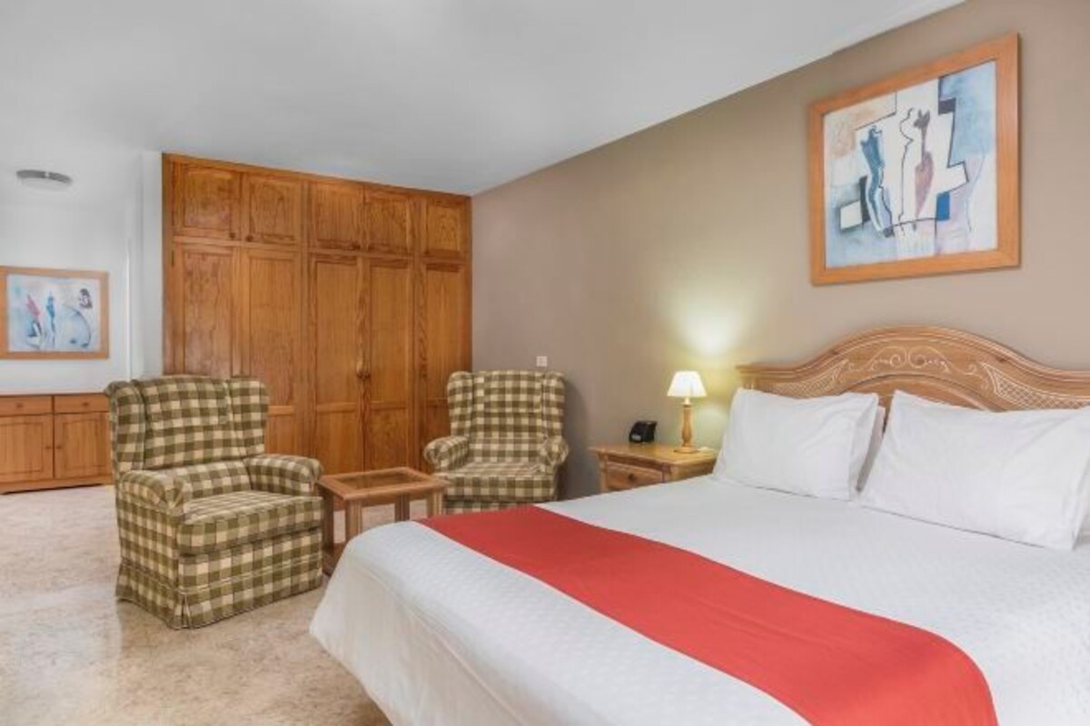 A hotel room with a large bed, two armchairs, a wooden wardrobe, and a painting on the wall.
