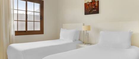 A hotel room with two single beds, a window with curtains, a bedside table with a lamp, and a painting on the wall.