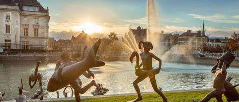 A sculpture of a swan and a child in a park with a fountain and buildings in the background.