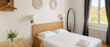 Habitación de hotel con cama de madera, dos lámparas fijadas a la pared, un espejo redondo y una ventana con cortinas.