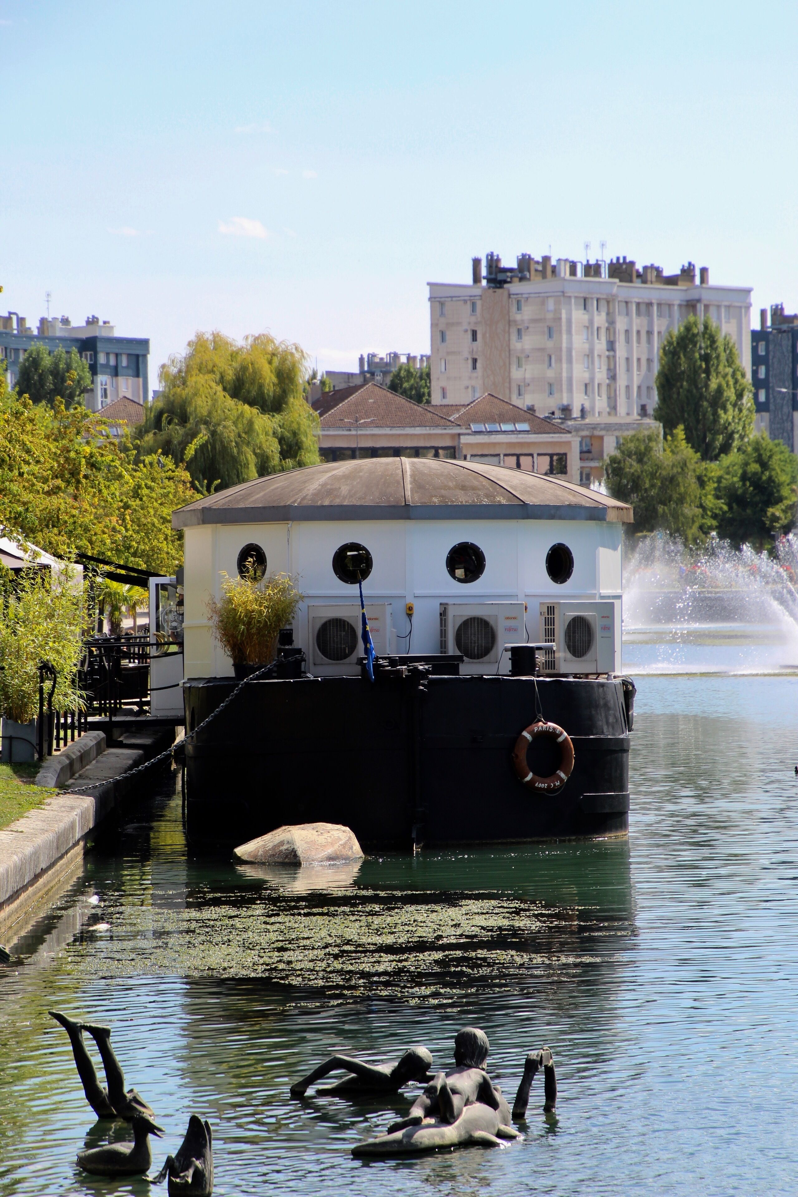 A boat with a white cabin and black hull, docked at a canal with water plants and ducks.