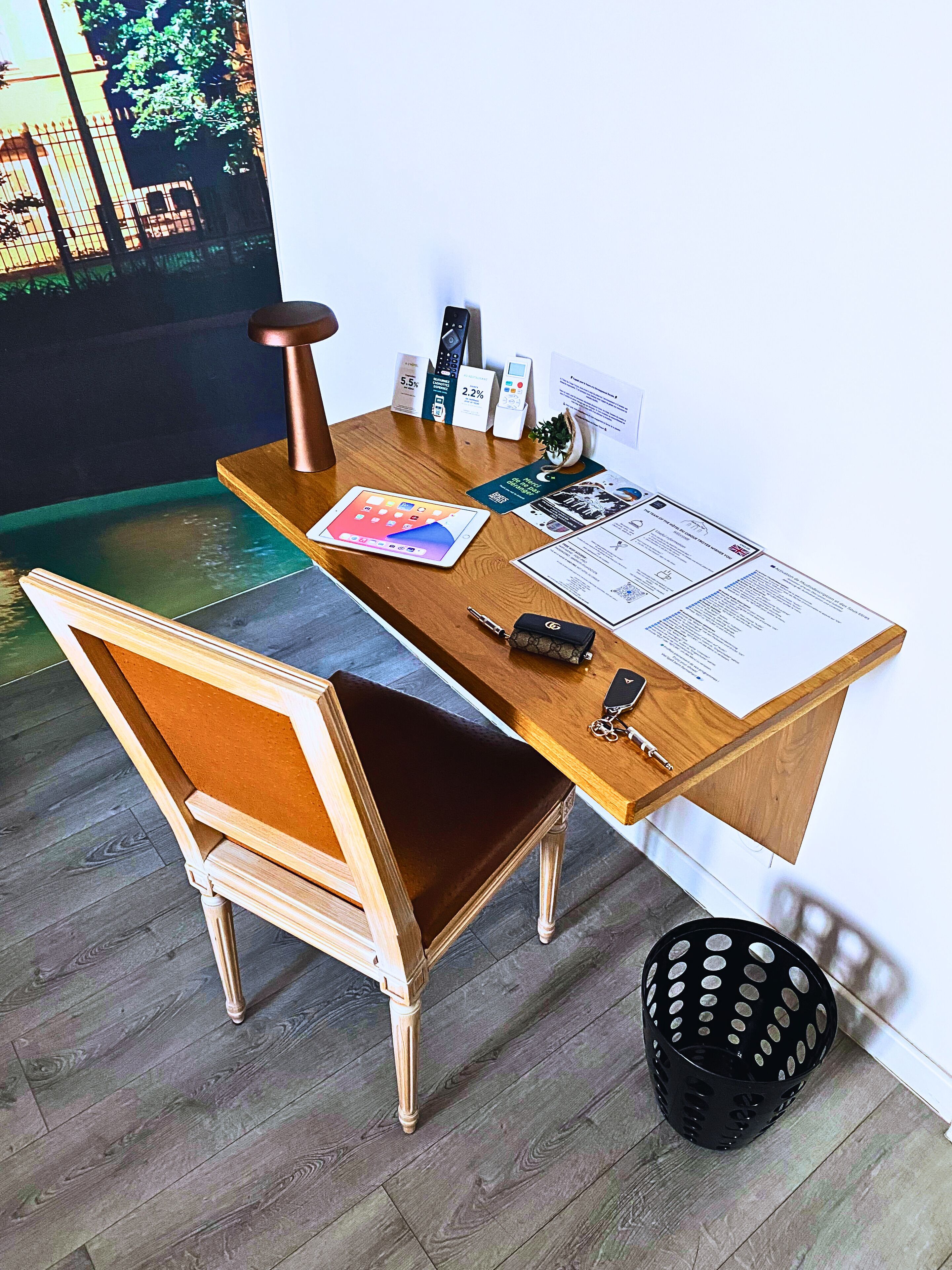 A wooden desk with a laptop, a smartphone, and a remote control. A chair with a cushion is positioned in front of the desk.
