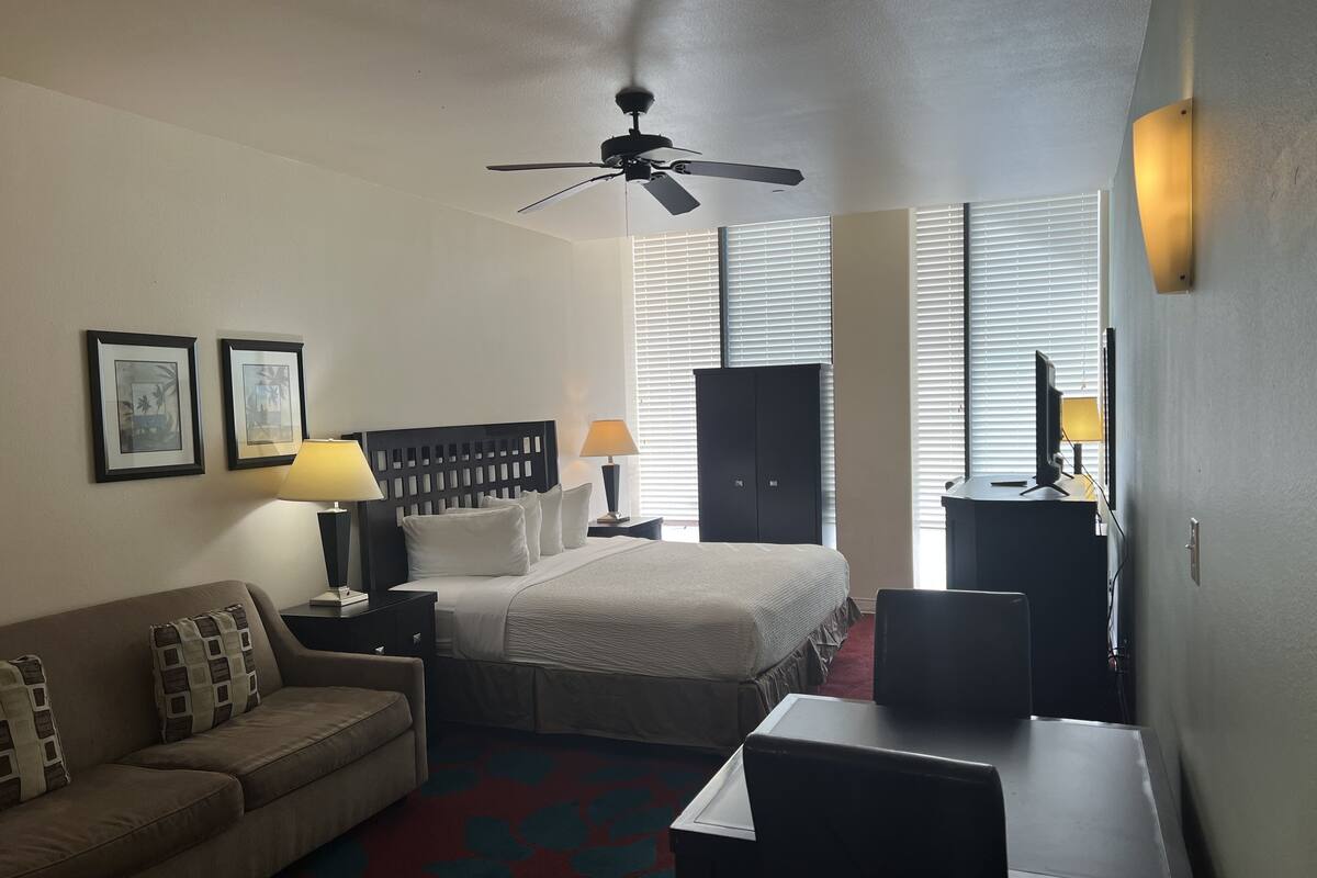 A hotel room with a bed, a desk, a chair, a sofa, a ceiling fan, and two wall-mounted lamps.