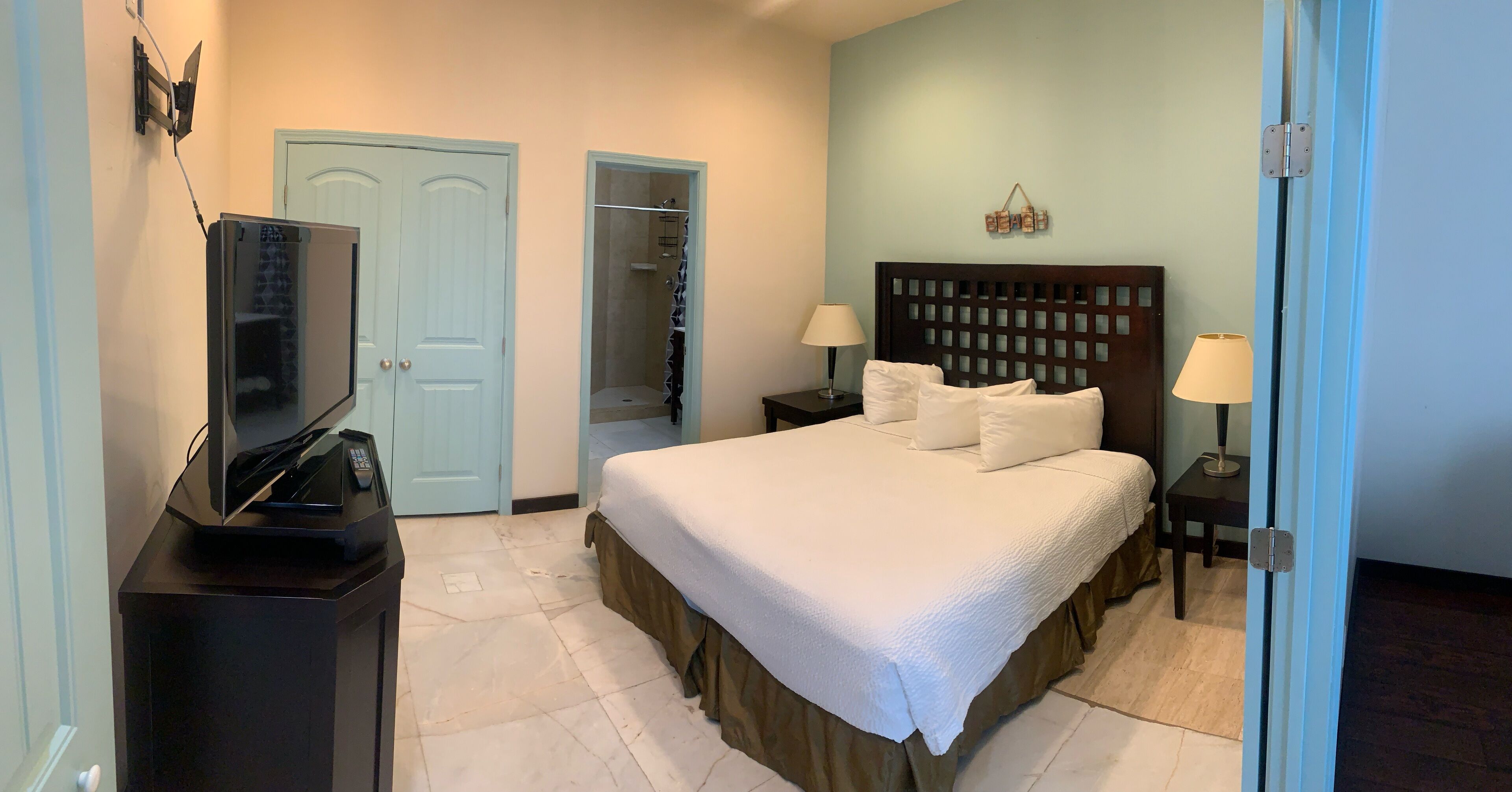 A hotel room with a bed, a TV on a stand, a nightstand with lamps, a closet, and a shower area.