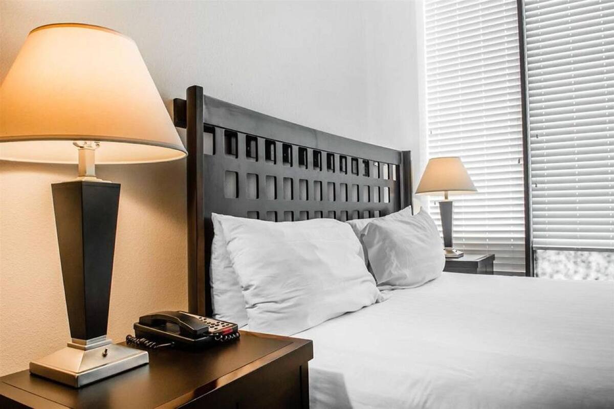 A hotel room with a bed, two bedside lamps, a telephone, and a window with blinds.