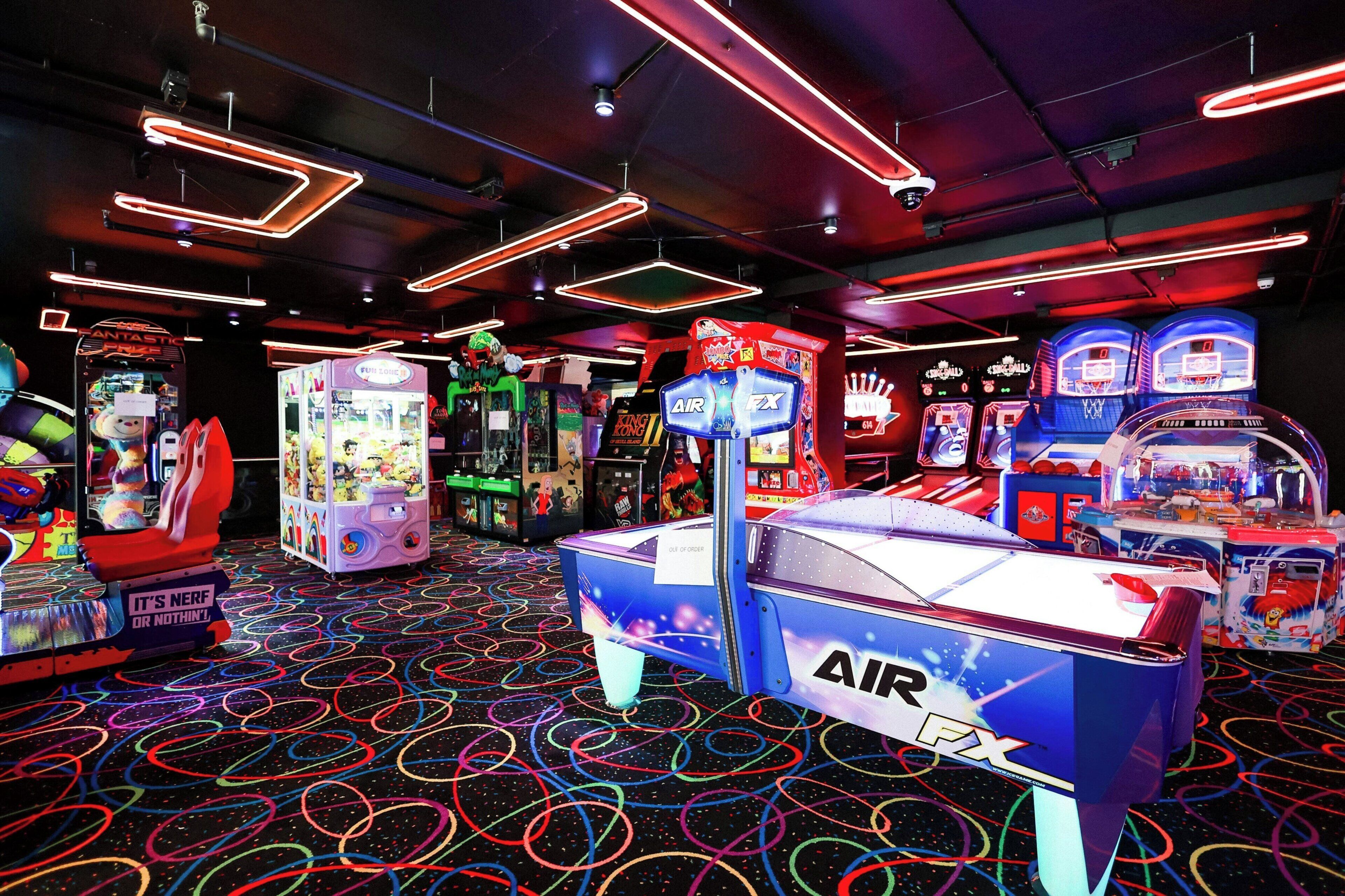 An indoor arcade with various gaming machines, neon lights, and a colorful floor pattern.