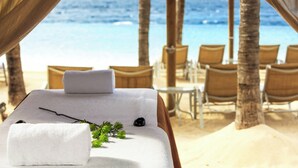 A massage table with a towel and a black stone, set on a beach with chairs and palm trees, overlooking the ocean. - Renaissance Wind Creek Curacao Resort (Willemstad)
