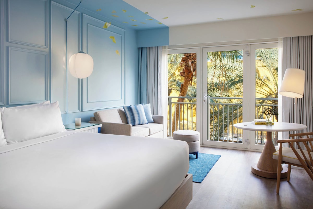 A hotel room with a large bed, a sofa, a small table, and a balcony with a view of palm trees.