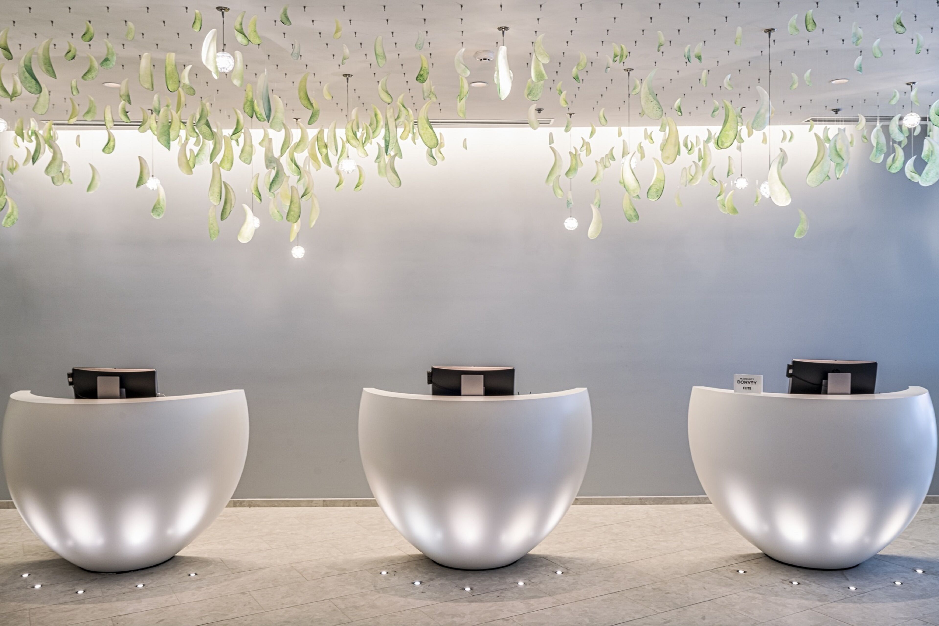 Un área de recepción con tres mostradores blancos y modernos, de forma redondeada, y un techo decorado con hojas verdes colgantes.