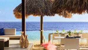 A beachside dining area with a view of the ocean, a thatched umbrella, and outdoor seating. - Renaissance Wind Creek Curacao Resort (Willemstad)