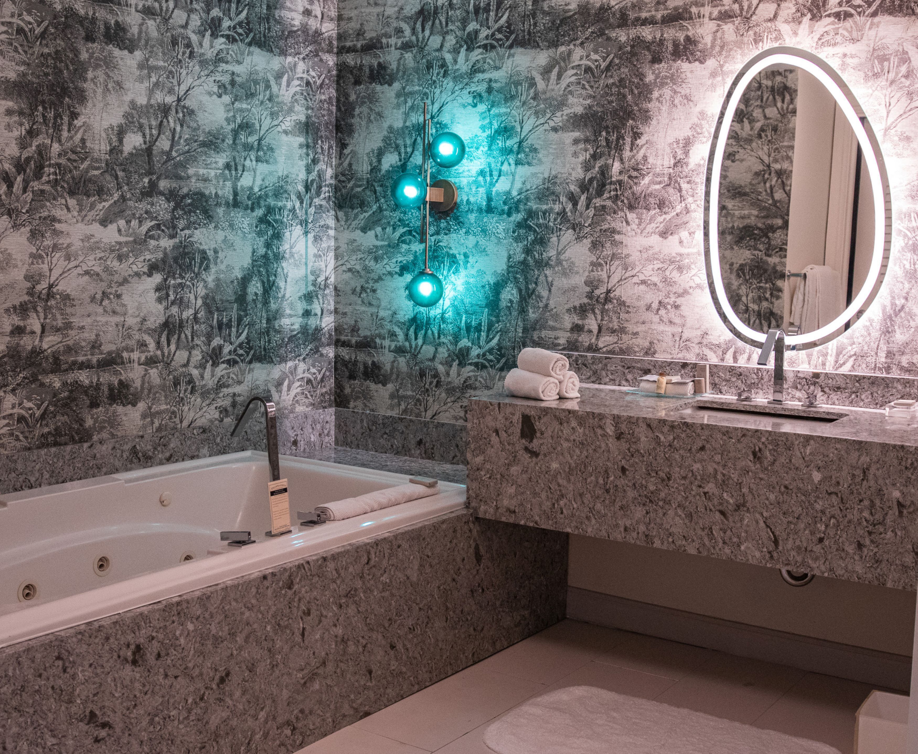 A bathroom with a bathtub, a marble countertop, a sink, a mirror, and a wall with a nature-themed wallpaper.