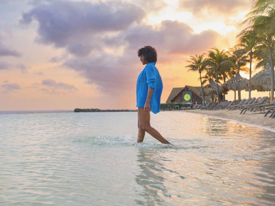 A person walking on a beach with a thatched-roof structure in the background.